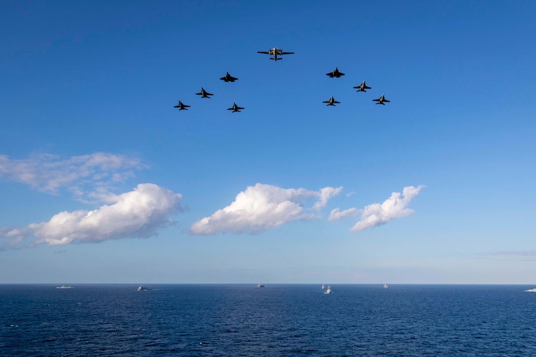 Aircraft fly in formation in a blue sky above a large group of ships in the ocean.