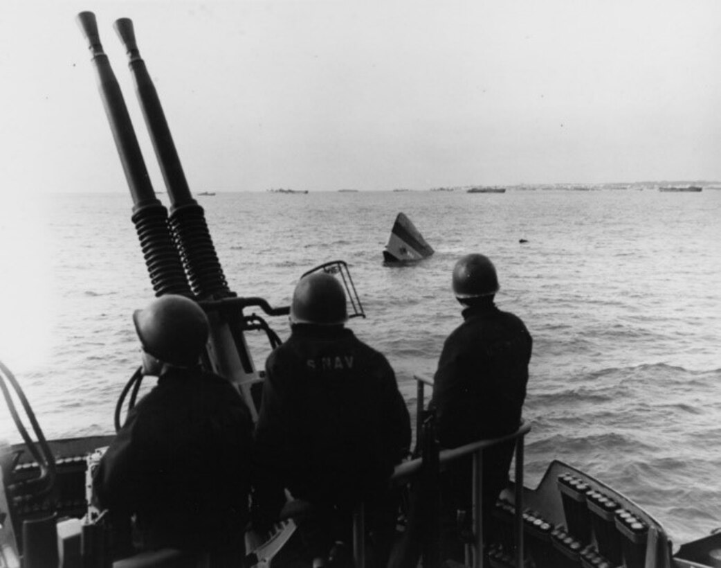 A crisp black-and-white photograph, taken from USS Brooklyn (CL-40), shows approximately fifteen feet of USS Portent’s (AM-106) bow sticking above the water on D-Day upon her sinking from a mine, 22 January 1944