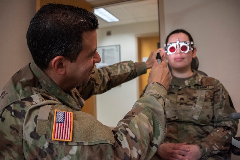 A uniformed service member adjusts vision testing equipment on another uniformed service member.