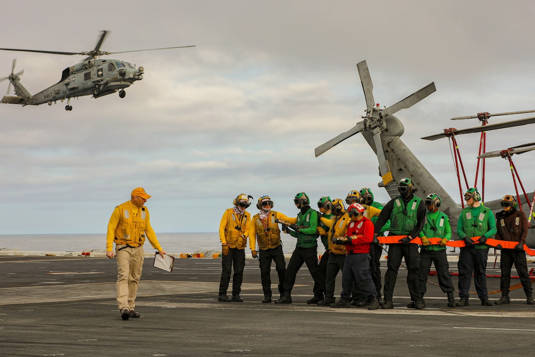 A sailor observes as a dozen sailors hold a red hose aboard a ship with the tail rotor of a helicopter in the background as airborne helicopter approaches to the left.