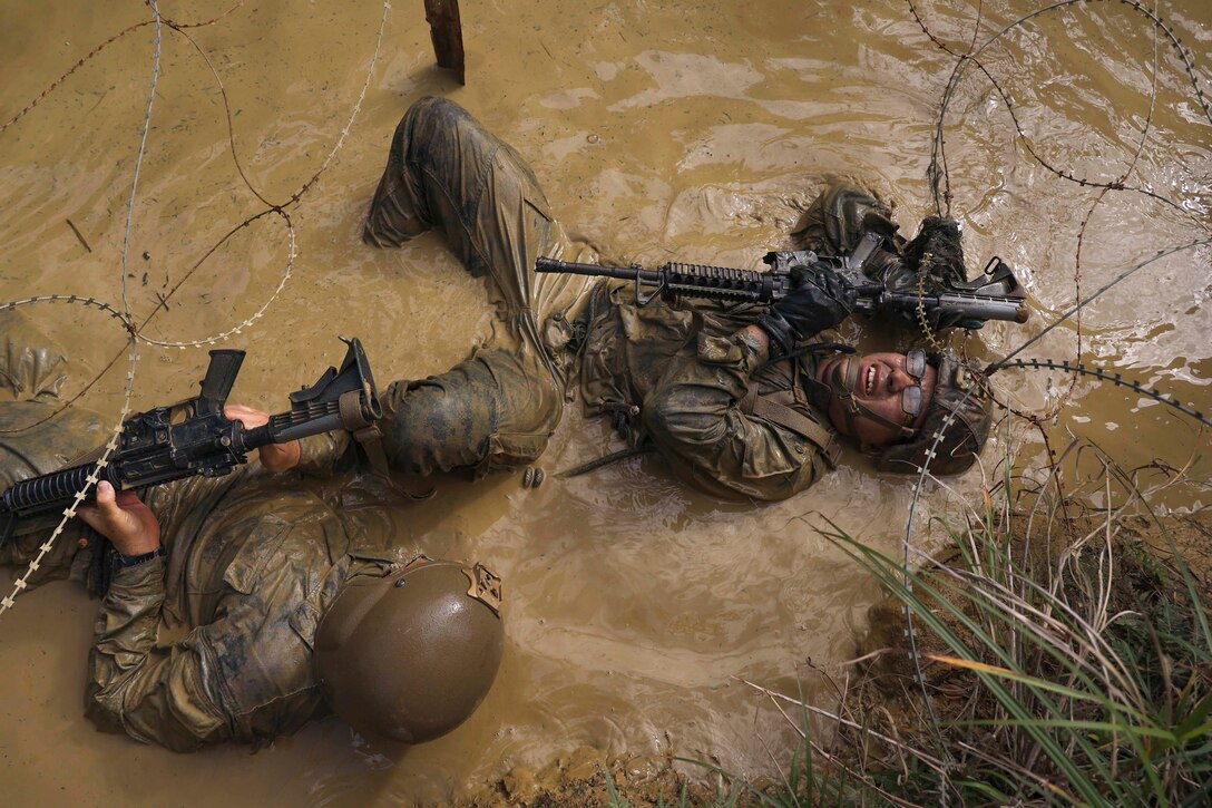 Marines in tactical gear lay upside down to move through mud under barbed wire.