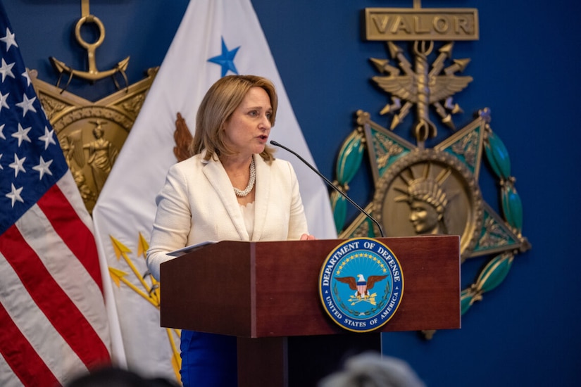 A woman stands at a lectern. The seal on the front of the lectern indicates she is at the Department of Defense.