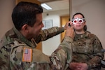 A uniformed service member adjusts vision testing equipment on another uniformed service member.
