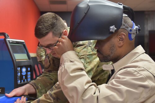 One person wearing a welding mask looks at a training tool while another person assists.