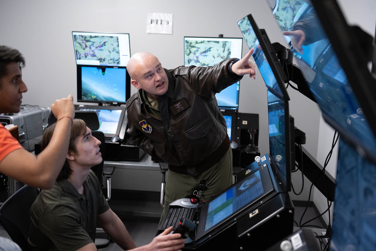 Two civilian students watch as a standing service member points to one of several monitor screens in a room.