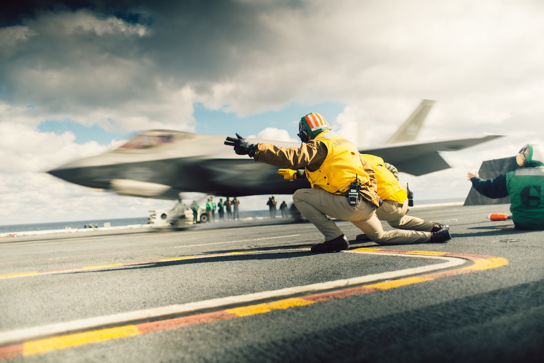 Two sailors kneel and point as a fighter jet prepares to launch from an aircraft carrier.