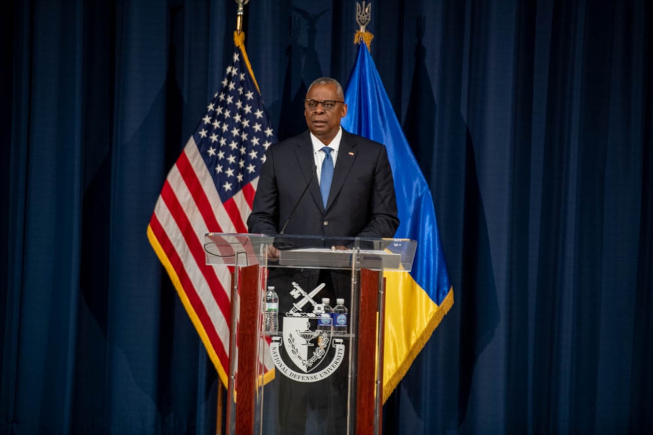 A man in a suit stands at a lectern with U.S. and Ukrainian flags in the background.