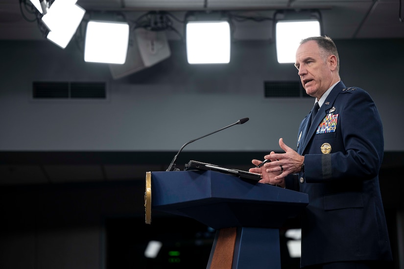 An Air Force major general stands at a podium while speaking into a microphone at a lectern beneath large ceiling lights.