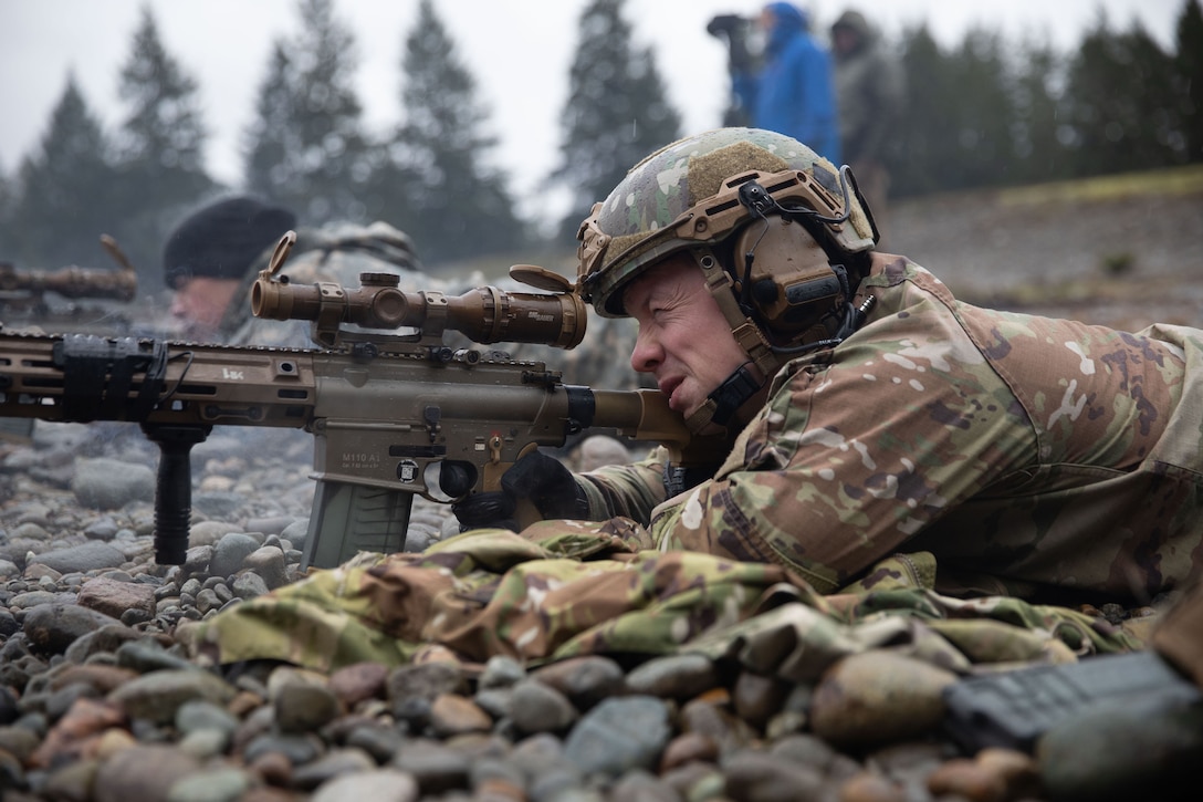 A soldier looks through a scope and fires a weapon while lying on a rock-covered ground.