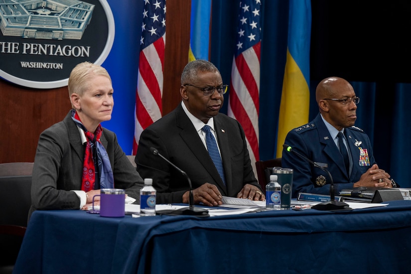 Three people are seated at a table with U.S. and Ukrainian flags in the background.