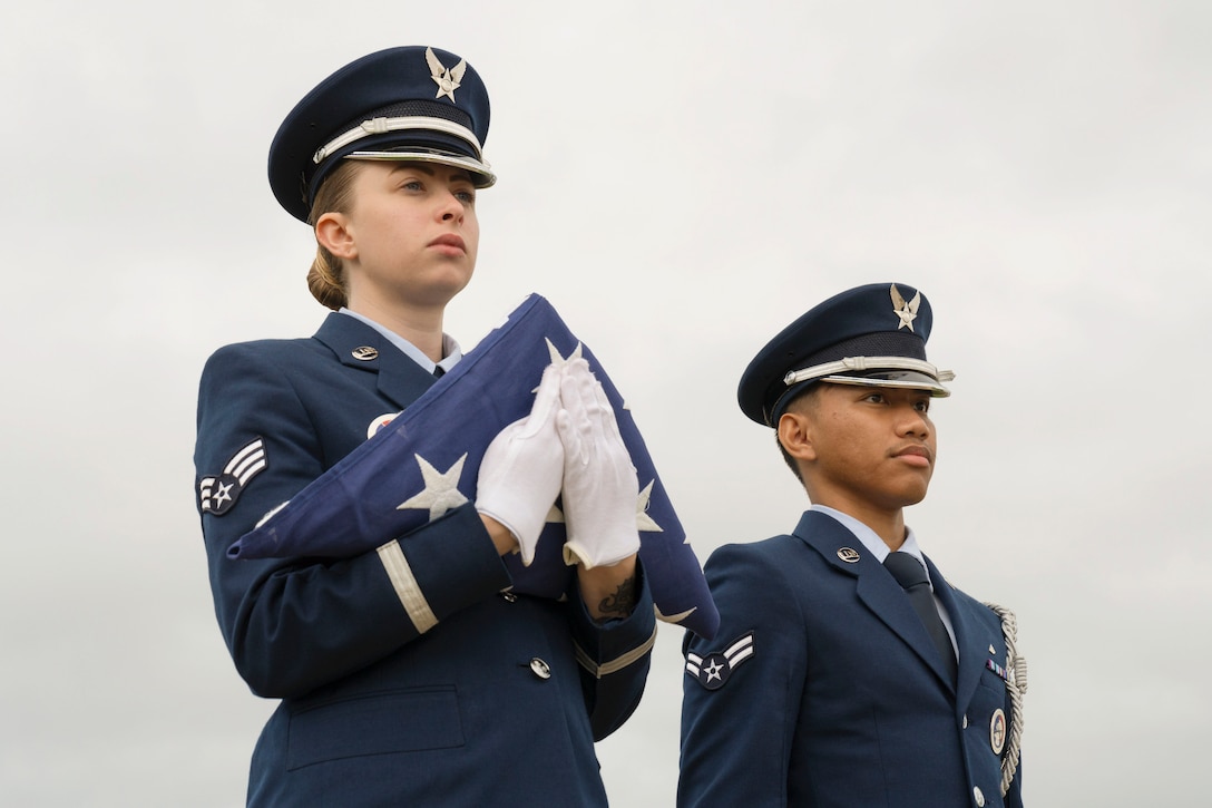 Two airmen in ceremonial uniform stand in formation as one holds a folded American flag.