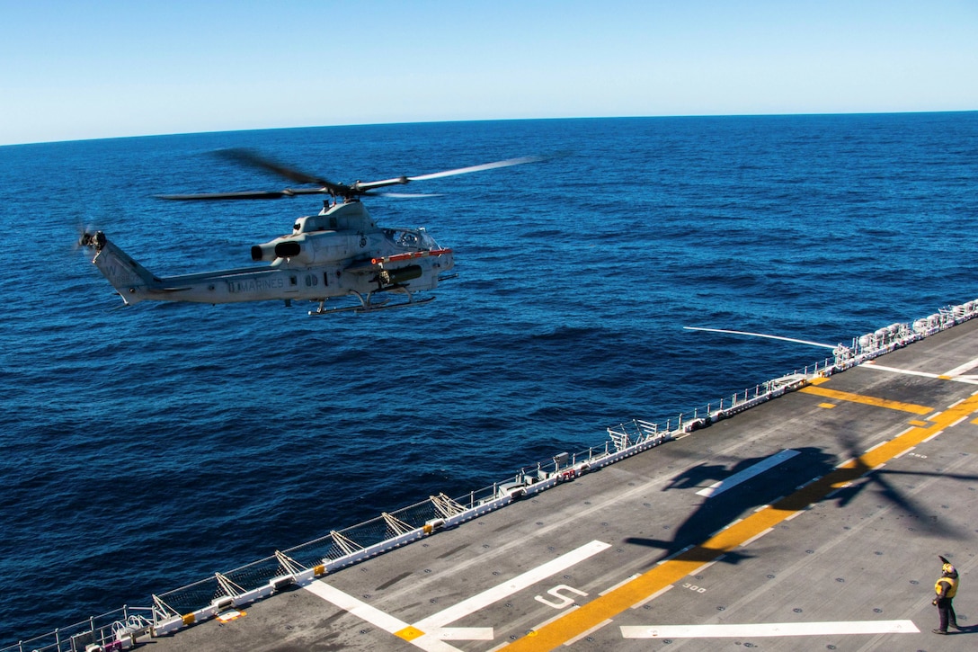 A sailor stands on a ship’s deck next to the shadow of a helicopter as it flies above.