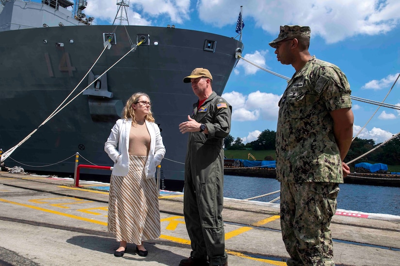 Two men in uniform speak with a woman. A military ship is behind them.