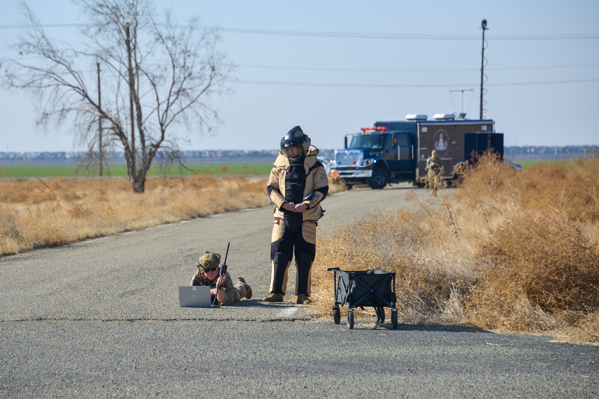 One Airman lies on his stomach on a road and uses a computer while the other stands in a bomb suit and waits for him.