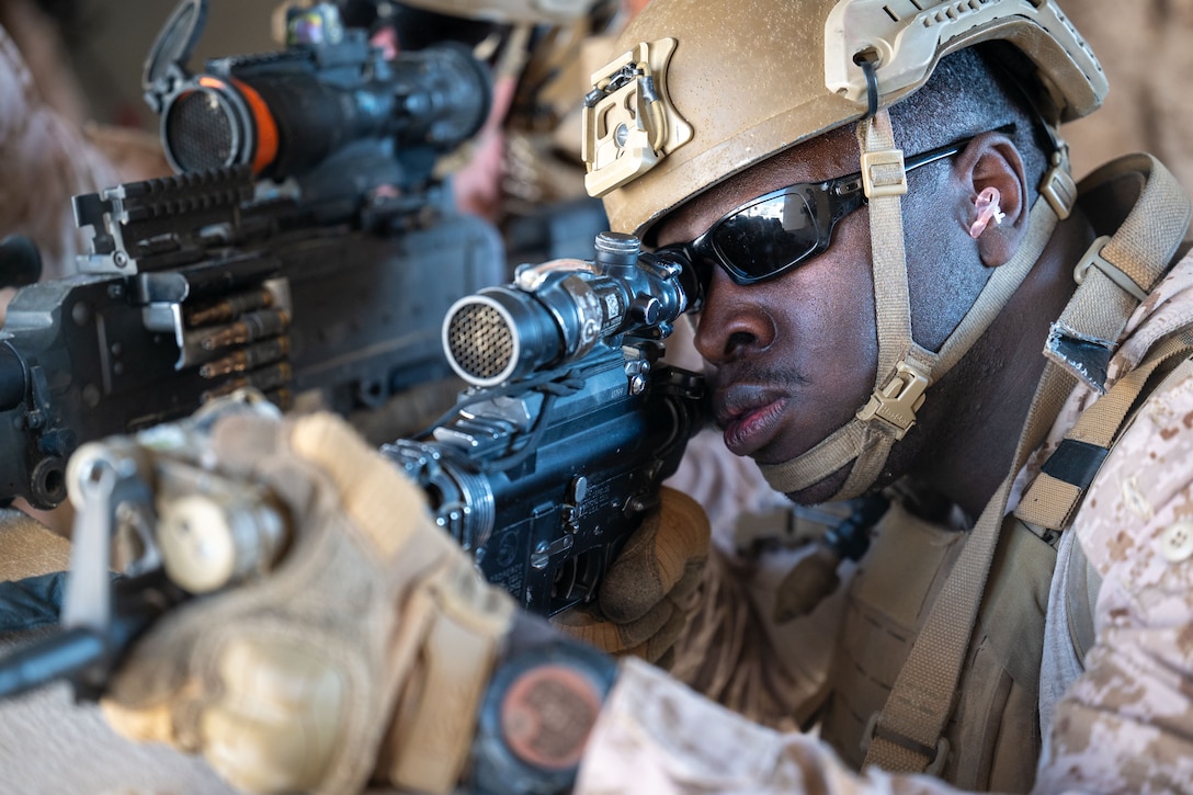 A Marine wearing dark sunglasses aims a weapon. Another Marine’s weapon can also be seen.