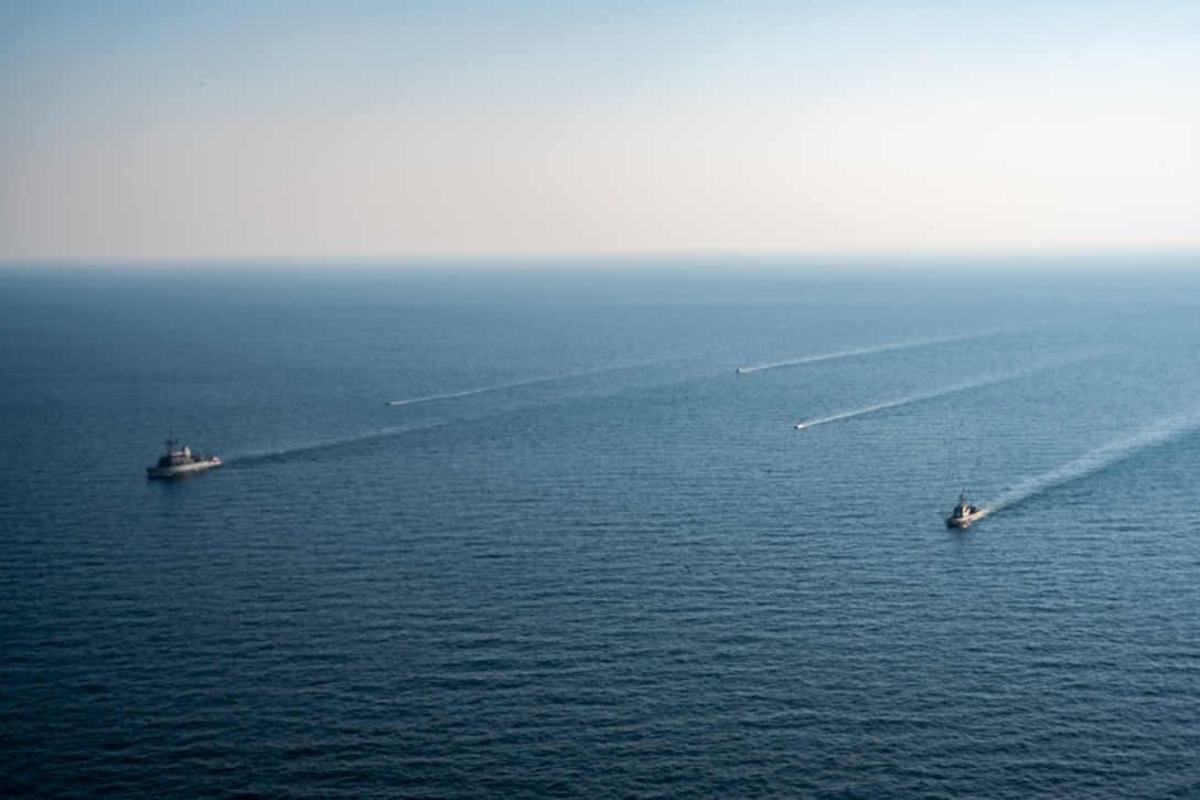 A group of ships and uncrewed vessels steam across a large body of water.