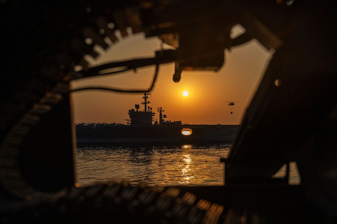 Distant view of a helicopter hovering over a ship, silhouetted against an orange sky, framed by equipment on a ship in the foreground.