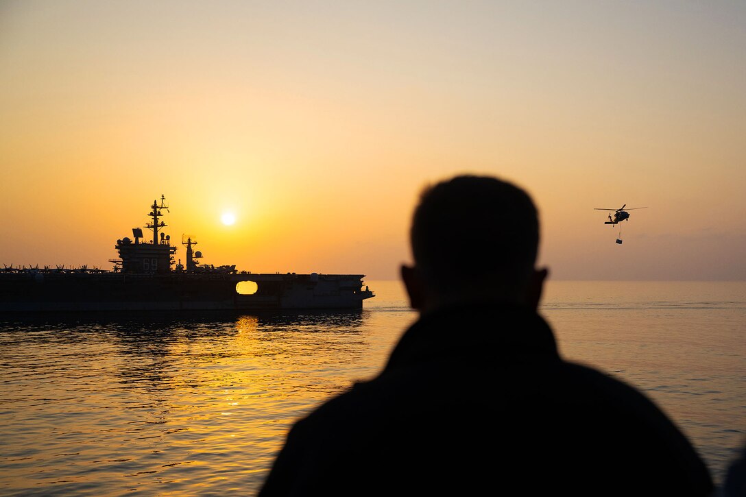 A sailor watches a helicopter flying near a ship in the distance, all silhouetted against an orange sky.