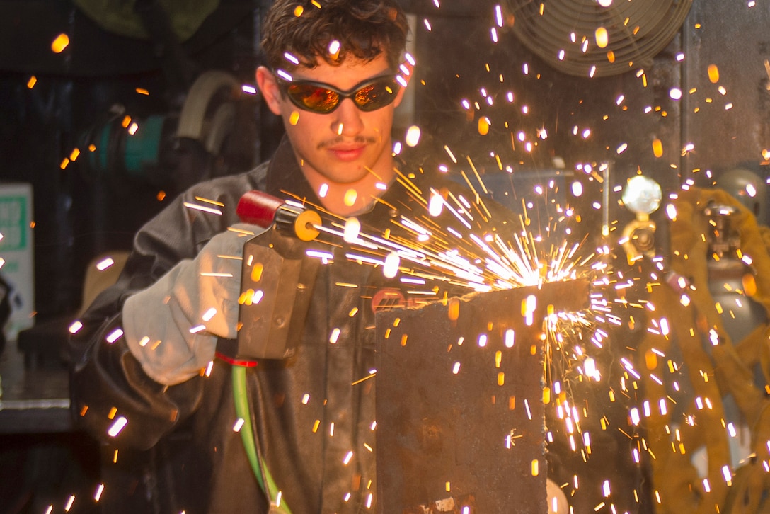 Sparks fly as a sailor wearing protective gear welds metal in a mechanical room.