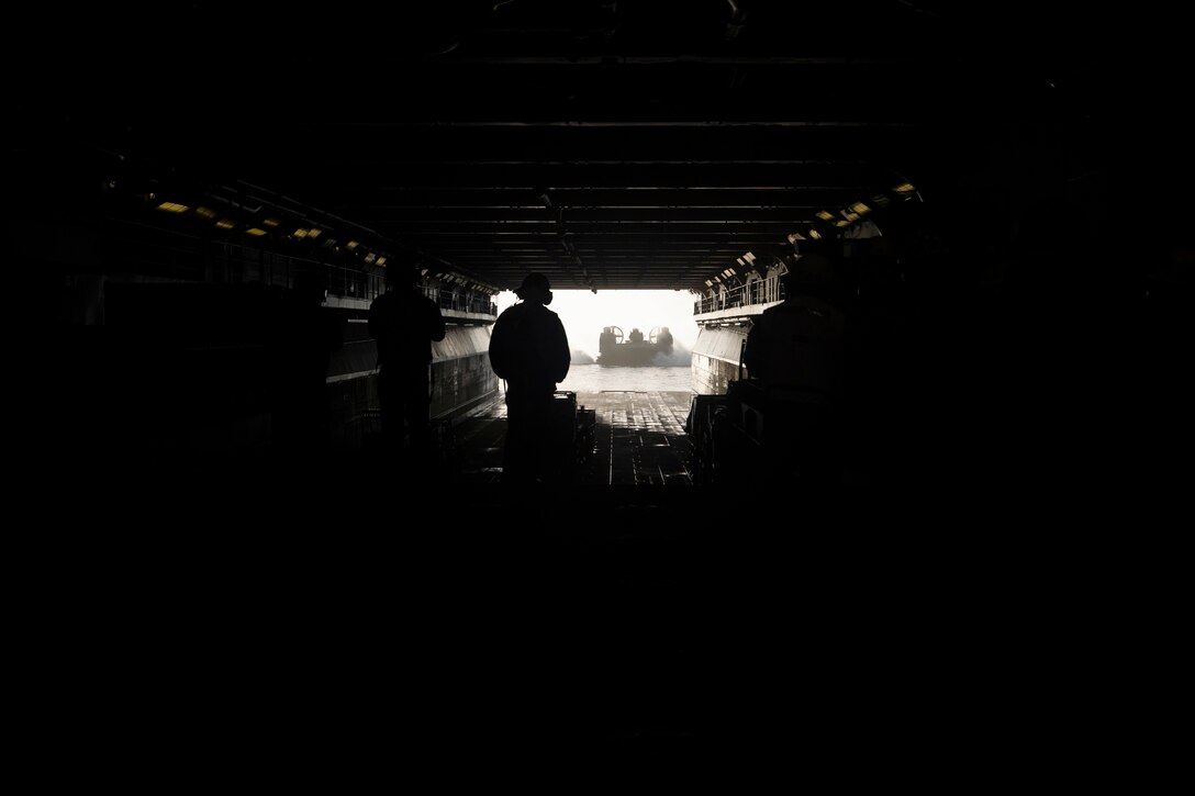 Sailors walk in a ship’s dark well deck toward an opening as a landing craft approaches.