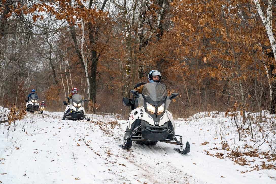 Four sailors ride snowmobiles on a snowy trail in the woods.