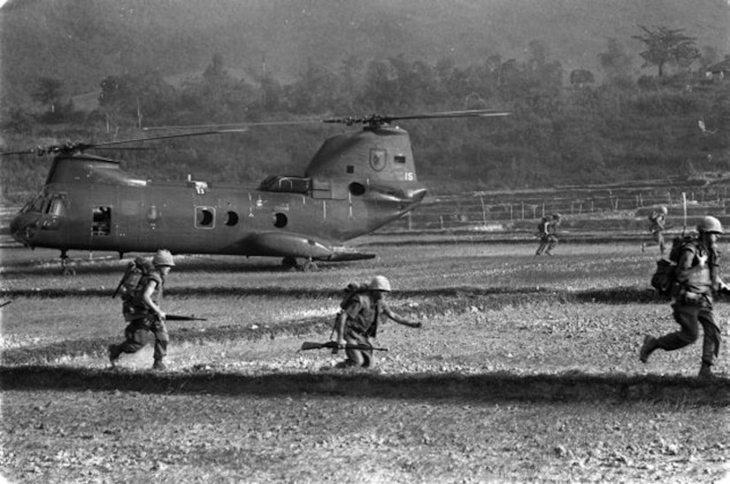 Service members with rifles race across a field, away from a helicopter.