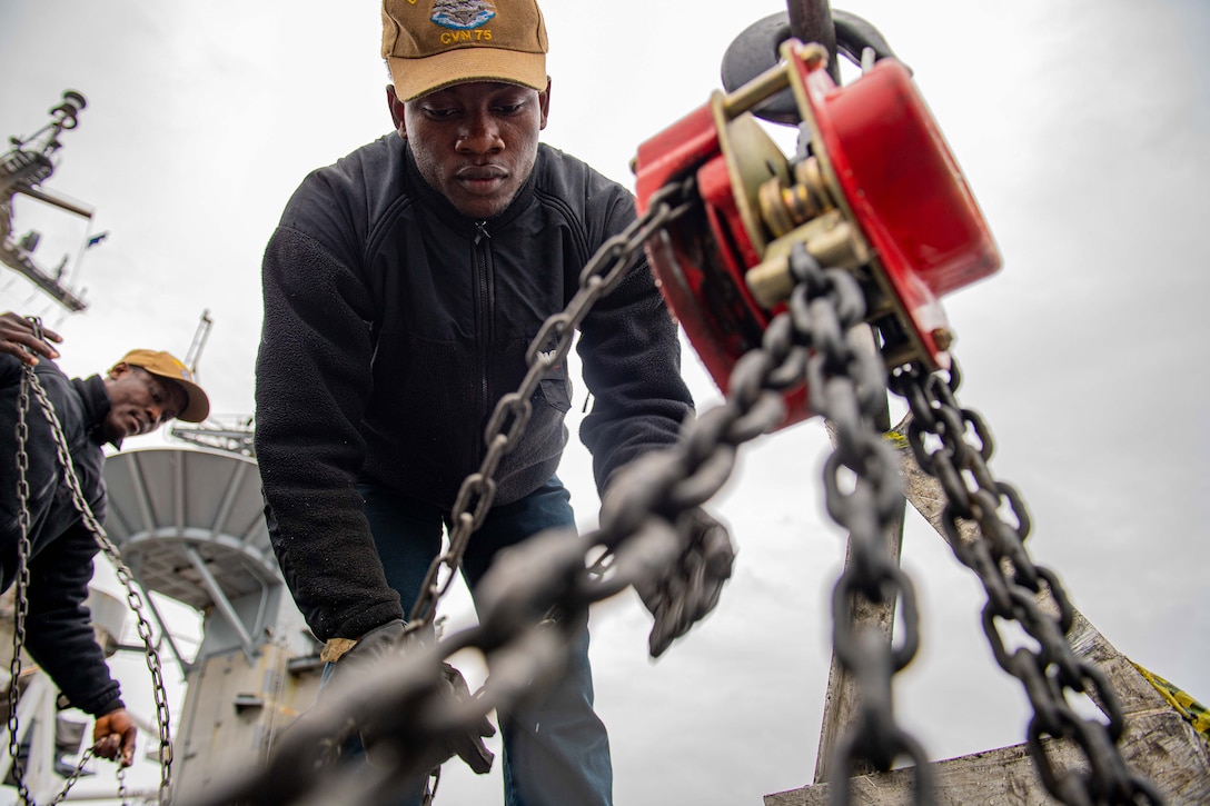 A sailor works on chains on a ship’s flight deck as another sailor works in the background.