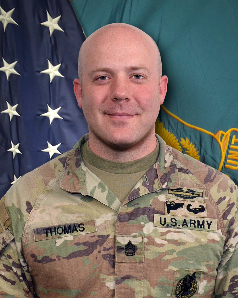 Man in U.S. Army uniform standing in front of two flags.