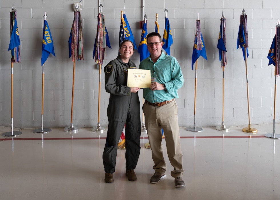 Two people pose with a certificate in front of flags