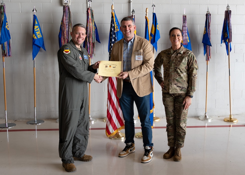 Three people pose with a certificate in front of flags