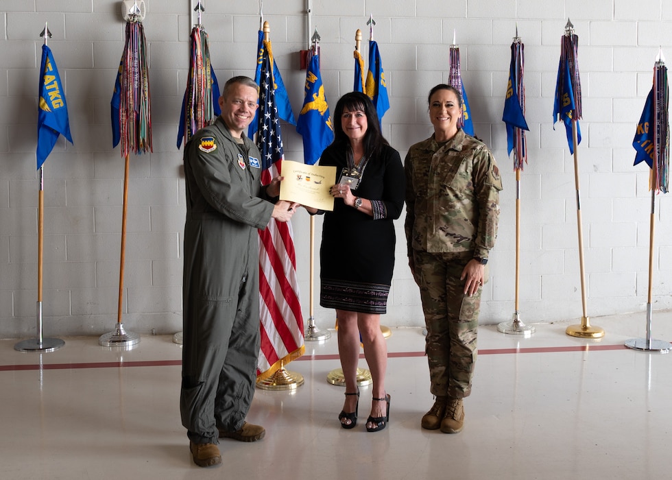 Three people pose with a certificate in front of flags