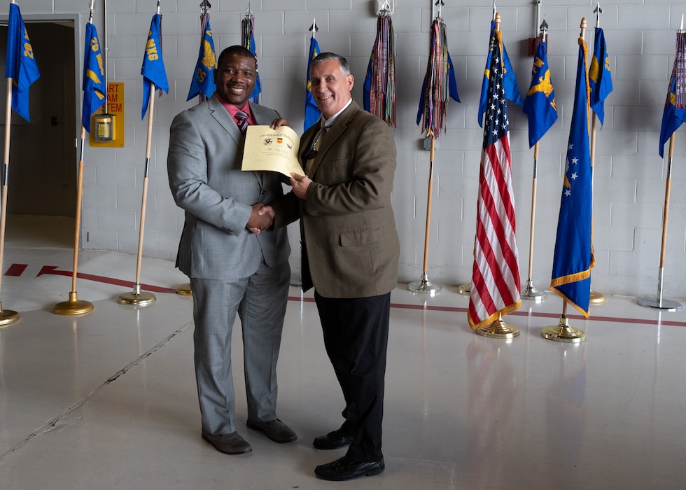 Two people pose with certificates in front of flags