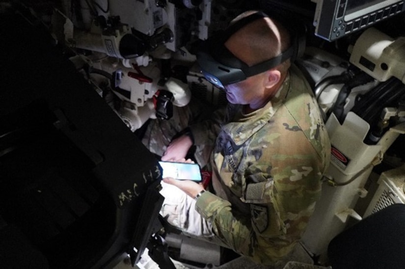 A soldier wearing a light headset holds a phone while seated around tech equipment.