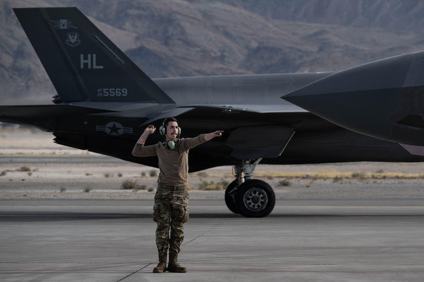 An airman gives signals while standing with an F-35A in the backdrop and hills beyond.