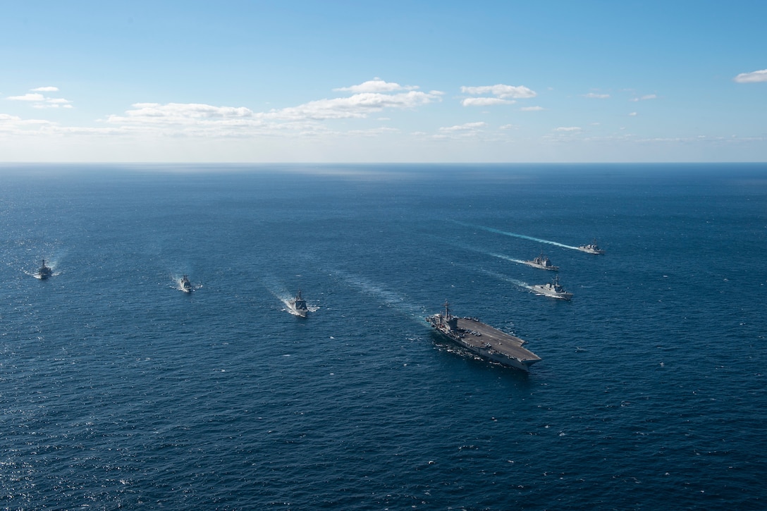 A group of military ships steam in formation through the water.