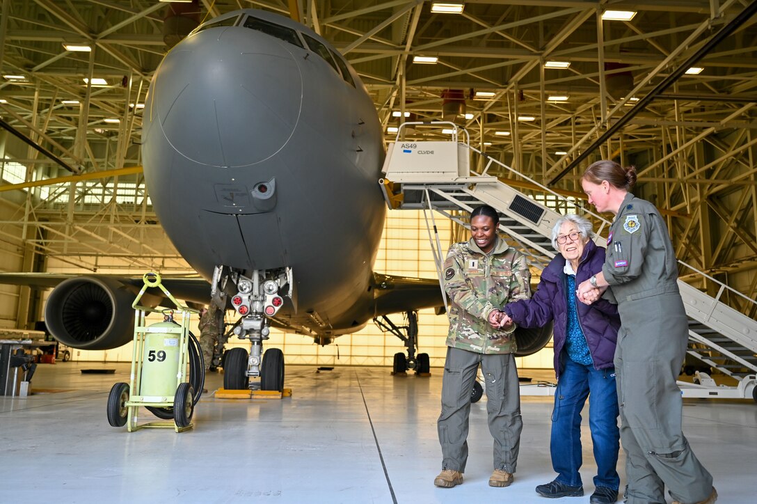 Two airmen walk with a World War II veteran in a hangar with a large aircraft in the background.