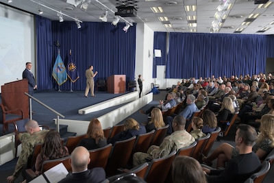 A Navy Captain in uniform and a civilian SES stand on the stage in front of a large audience