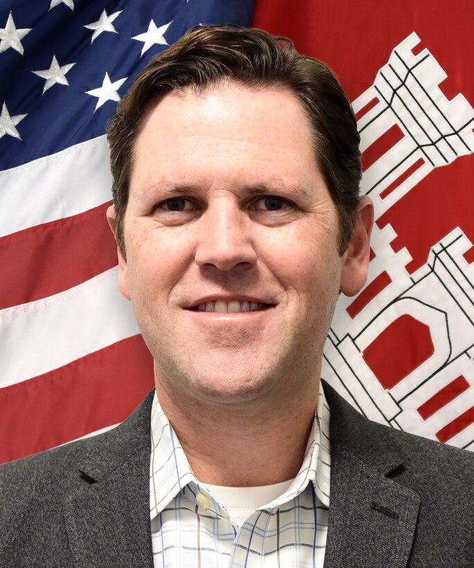 Head and shoulders shot of white male standing in front of red, white and blue flag, and a red and white flag.