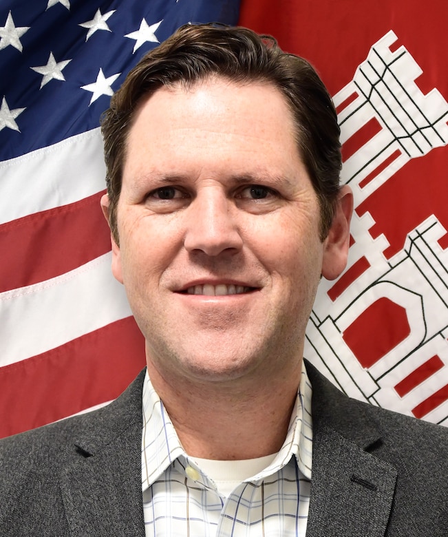 Head and shoulders shot of white male standing in front of red, white and blue flag, and a red and white flag.