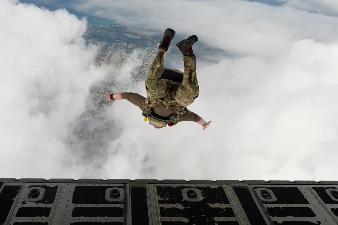 An airman free falls near clouds with the aerial view of a city in the background and an open door for an aircraft in the foreground.