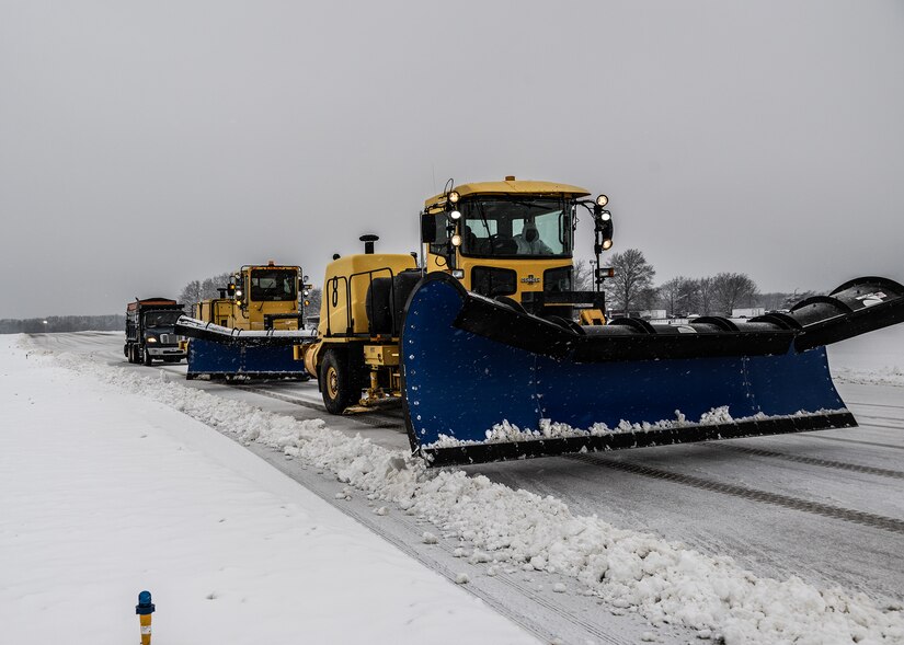 Three yellow snow plows drive on flight line.