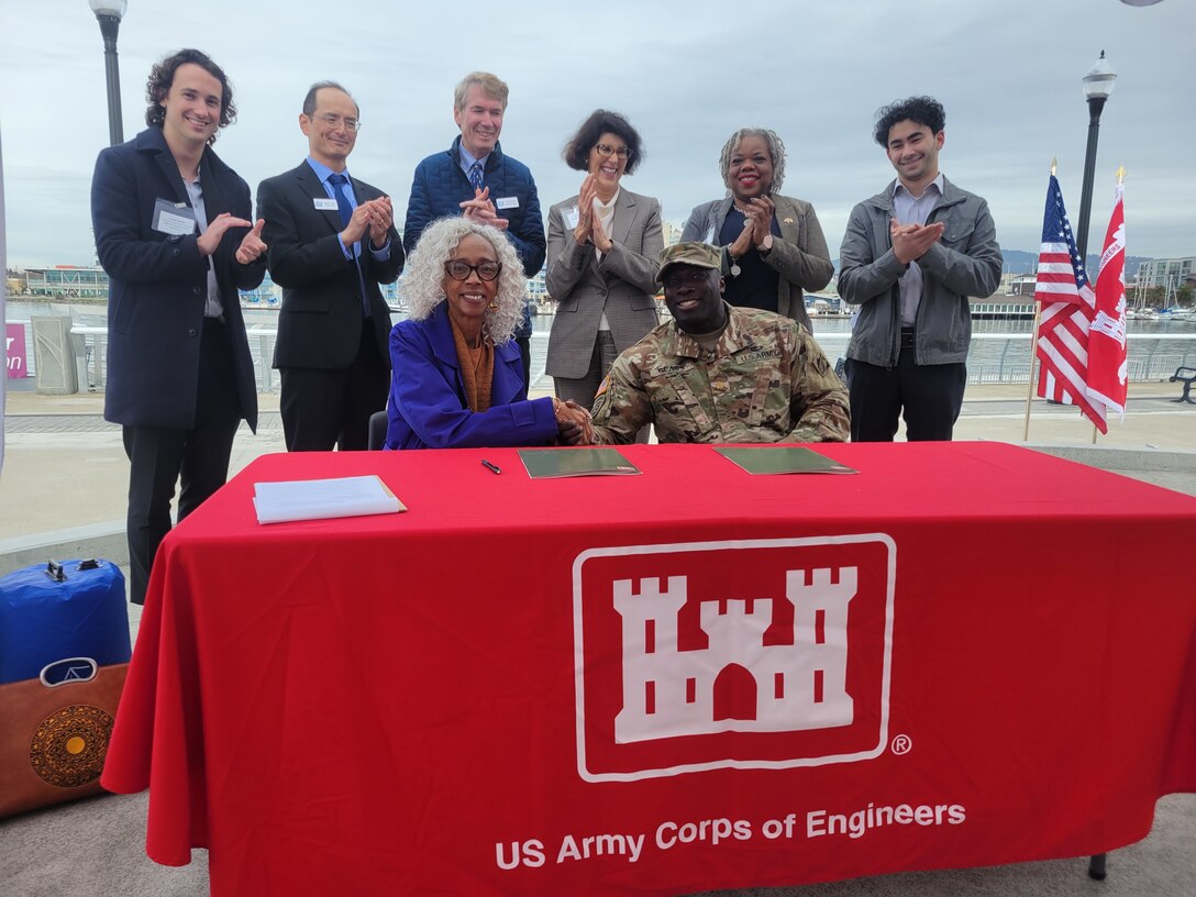 Six men and women stand behind one black male and female shaking hands while sitting at a table covered in a red and white castle tablecloth.