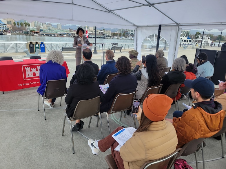 A white female with dark hair in a gray suit stands in front of a group of people sitting, as she speaks about the recycled water project.