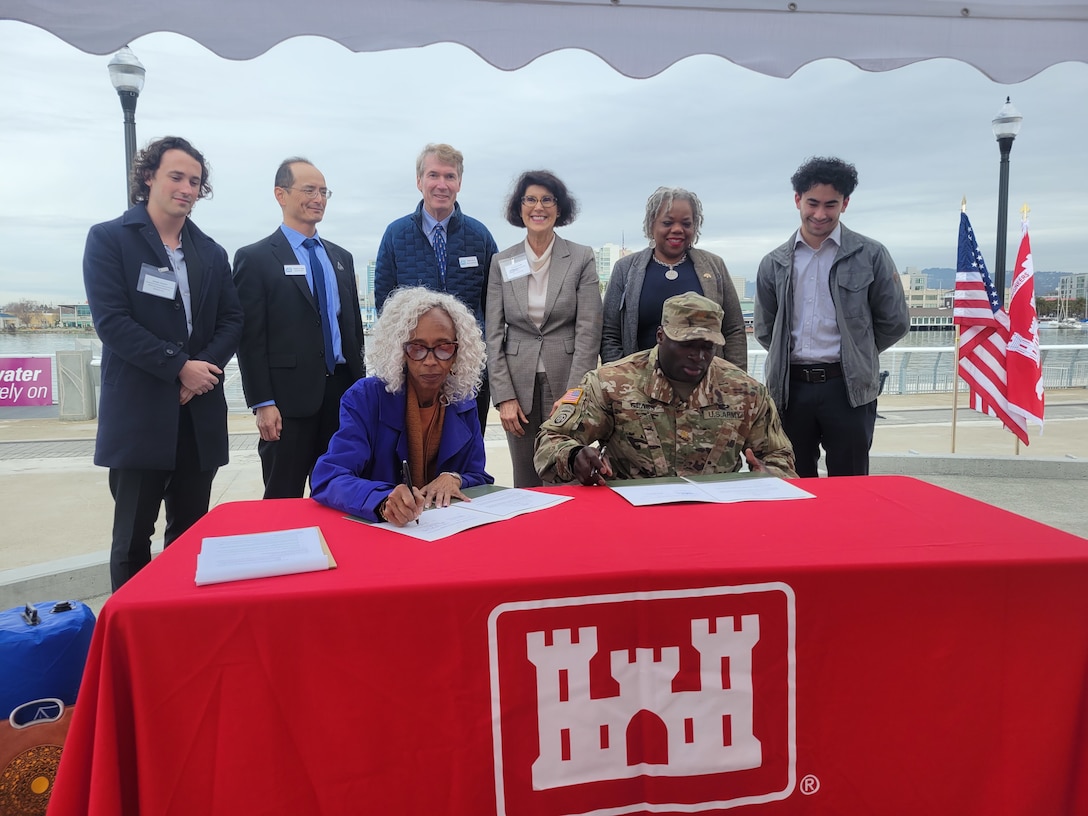 Six men and women stand behind two seated men and women who are signing documents on a table covered in a red tablecloth.