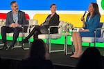 A person speaks into a microphone as two other people listen. All three are sitting on a stage.