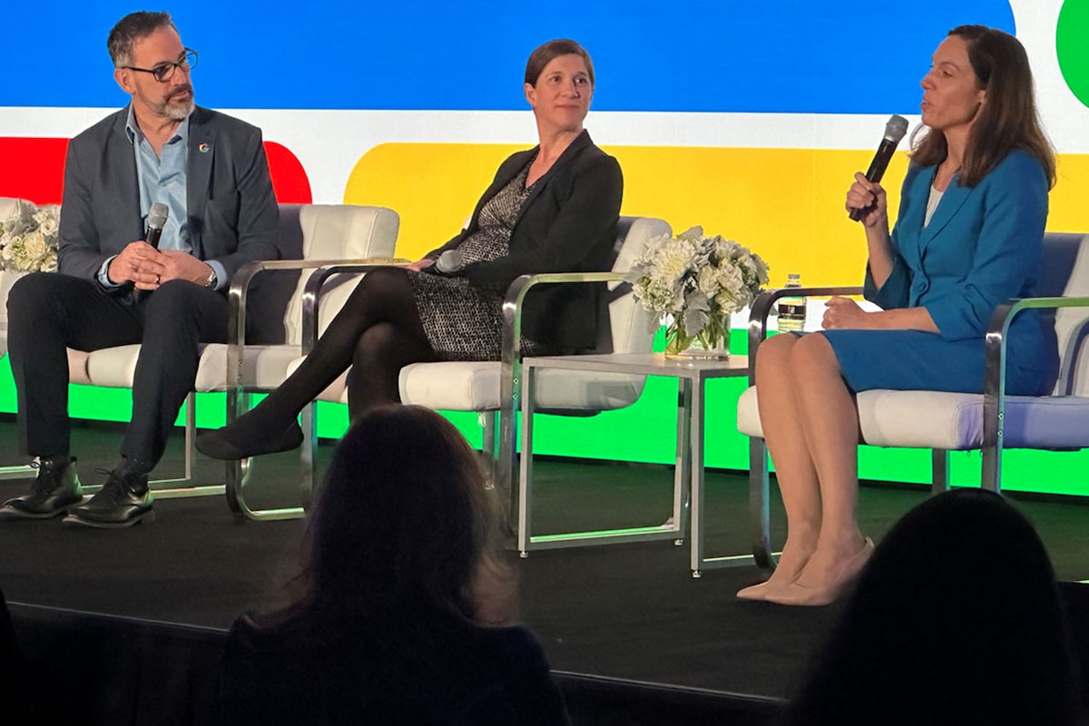 A person speaks into a microphone as two other people listen. All three are sitting on a stage.