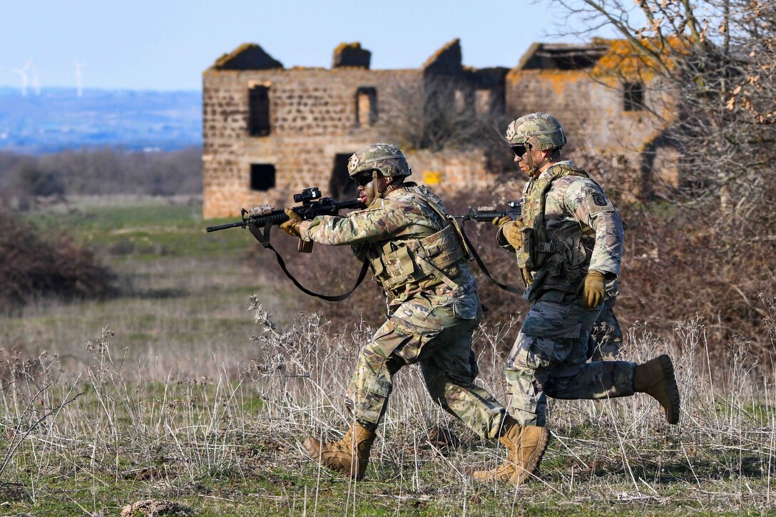 Two soldiers carrying weapons run through a field with a burned-out house in the background.