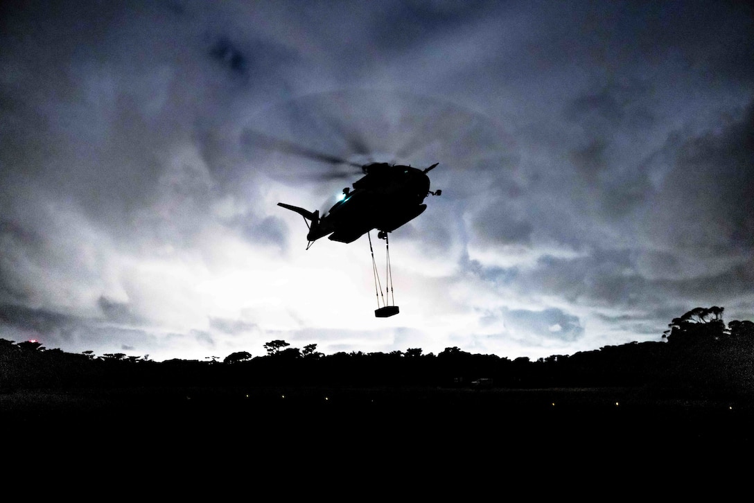 A helicopter lifts a pallet from the ground against a darkish sky.
