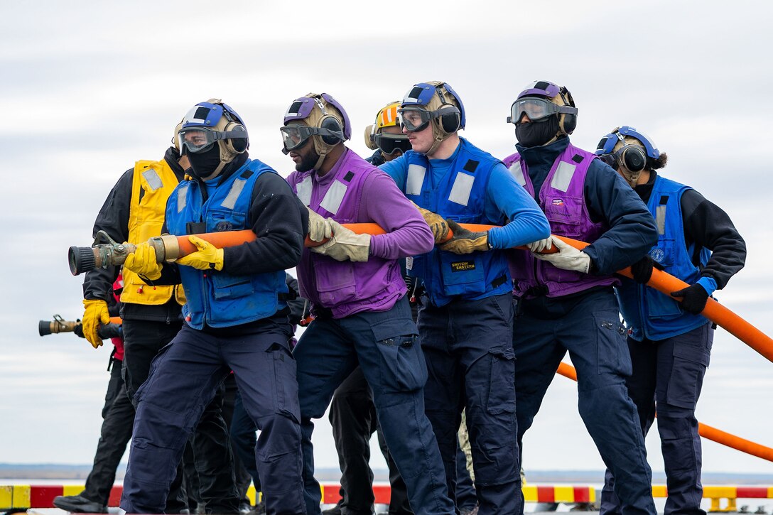 A group of sailors hold a hose on the flight deck of a Navy ship.