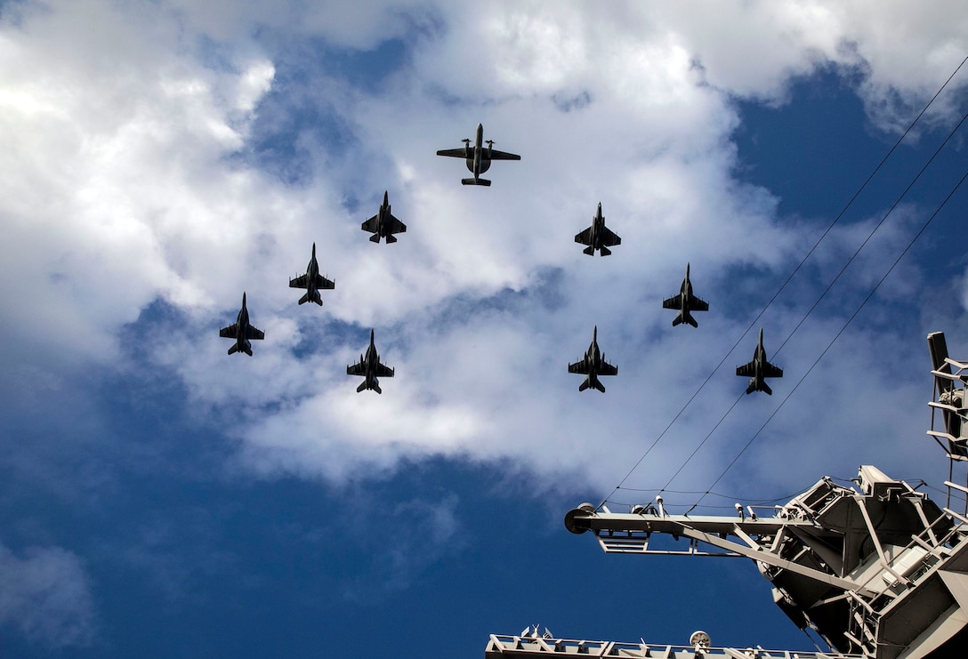 Military aircraft fly in formation over a Navy ship.
